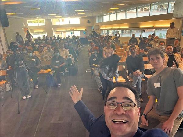 A dark haired man dressed in a sport coat and tshirt, 笑, takes a selfie with an amber-t一个d roomful of people behind him.